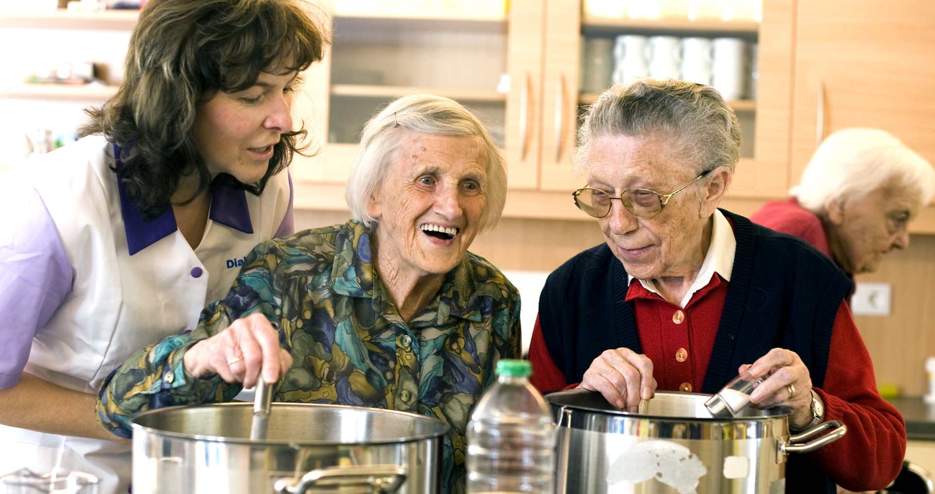 Zwei ältere Frauen und eine Pflegekraft stehen in einer Küche und lachen.
