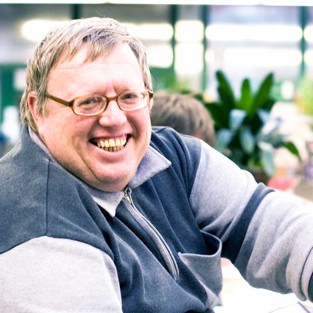 Ein Mann mit einer Brille lacht in die Kamera. Er arbeitet in einer Werkstatt.